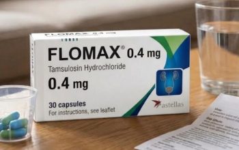 A close-up photograph of a box of Flomax 0.4 mg tablets, a glass of water, a medical pill cup with blue and green pills, and a patient information leaflet on a wooden coffee table in a living room.